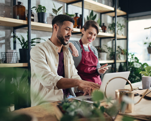 man and woman working together in a bright indoor garden setting with plants discussing ideas while using a laptop